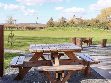 A wooden picnic table on a gravel area with a fire pit and log stools in a grassy field at Bouquet in Little Kelk near Bridlington