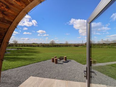 A view of a grassy field with tree stumps around a fire pit seen from a wooden structure with an open glass door at Bevy in Little Kelk near Bridlington