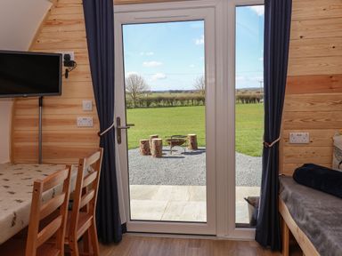 A room with wooden walls a table and chairs a mounted TV and a glass door leading to a grassy outdoor area with tree stumps around a fire pit at Bevy in Little Kelk near Bridlington