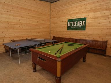A game room with a pool table and a table tennis table with wooden benches and a sign reading Little Kelk Glamping Site