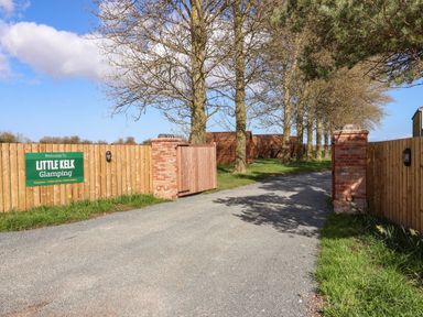 An entrance gate with brick pillars and wooden fencing along a driveway with trees at Little Kelk Glamping