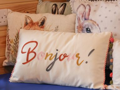 A white pillow with the word Bonjour embroidered in various colors placed among animal-themed cushions on a blue surface