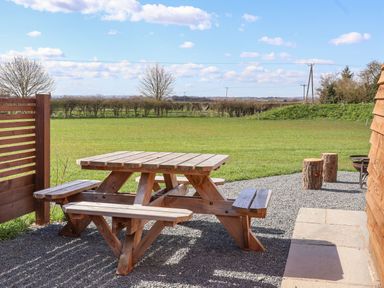 A wooden picnic table with benches on a gravel area next to a wooden fence and building with grass field and trees in the background at Husk in Little Kelk near Bridlington