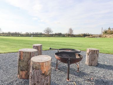 A fire pit with a grill and tree stump seats on gravel with a grassy field and goalposts in the background at Husk in Little Kelk near Bridlington