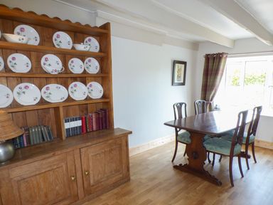 A dining room with a wooden table and chairs and a wooden cabinet with plates and books at Spring Garden Cottage in Laugharne