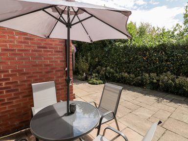 An outdoor patio area with a round table under a large umbrella and three chairs near a brick wall and bushes