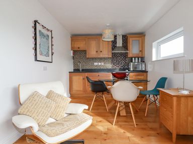 A kitchen with wood cabinets a glass dining table with three chairs a white armchair with cushions and a wooden side table with lamp at Lapwing in Perranporth