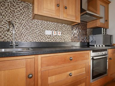 A kitchen countertop with a sink, wooden cabinets, a built-in oven, a range hood, a microwave, and a tiled backsplash at Lapwing in Perranporth