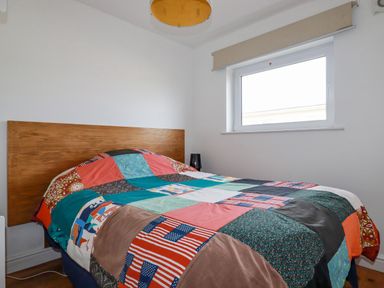 A bedroom with a wooden headboard a patchwork quilt on the bed a window with a beige blind and a black lamp on the floor at Lapwing in Perranporth