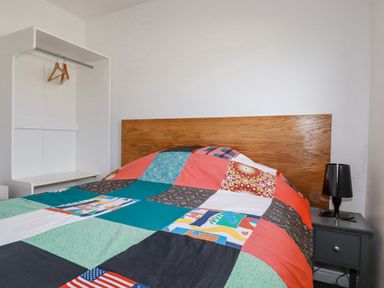 A bedroom with a bed covered by a colorful patchwork quilt a wooden headboard a small black side table with a black lamp and a white open closet with hangers at Lapwing in Perranporth