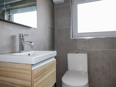 A bathroom with a white sink on a wooden cabinet a toilet a window and gray tiled walls at Lapwing in Perranporth