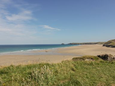 A sandy beach with people and grassy foreground with cliffs in the background at Lapwing in Perranporth