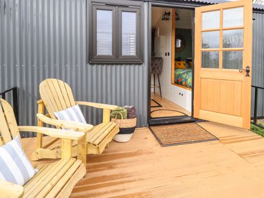 An outdoor wooden deck with two wooden chairs with cushions a basket with blankets an open wooden door leading to an interior space at Portland Luxury Glamping in Nottingham