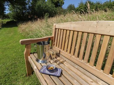 A wooden bench on grass with a metal ice bucket holding a bottle and two glasses with drinks and a plate with olives at Portland Luxury Glamping Nottingham