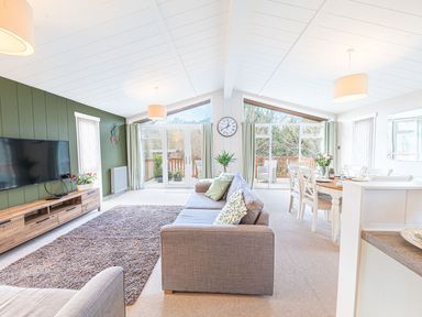 A living room with a grey sofa and a dining table with chairs near large windows with green curtains at Fern Lodge in Allithwaite near Cartmel
