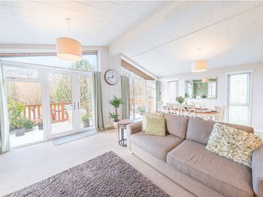 A living room with a sofa dining table plants and large windows at Fern Lodge in Allithwaite near Cartmel