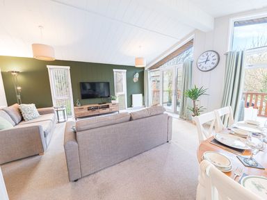 A living room with two sofas a television on a stand wall clock dining table set with plates and glasses at Fern Lodge in Allithwaite near Cartmel