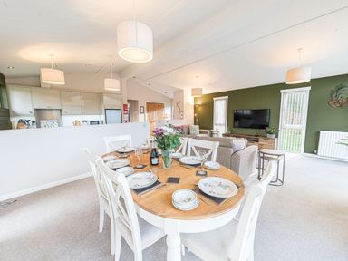 A dining table set with plates glasses and cutlery beside a kitchen and living room area at Fern Lodge in Allithwaite near Cartmel