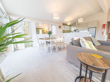 An open plan living room and dining area with a table and chairs a sofa with cushions and a small wooden side table with books at Fern Lodge in Allithwaite near Cartmel