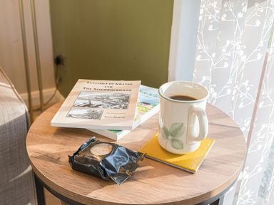 A round wooden table with a mug of coffee on a yellow coaster two books and a wrapped biscuit at Fern Lodge in Allithwaite near Cartmel