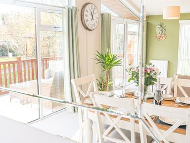A dining area with a wooden table set with plates glasses and a vase of flowers and white chairs near large windows with green curtains at Fern Lodge in Allithwaite near Cartmel