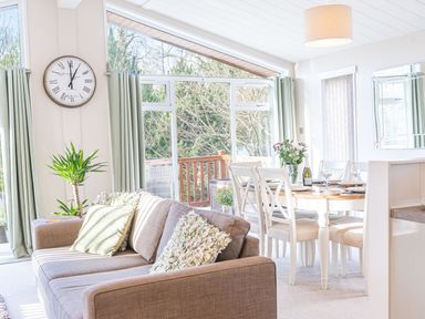 A living room with a sofa pillows a large clock green curtains and a dining table set with chairs and flowers at Fern Lodge Allithwaite near Cartmel