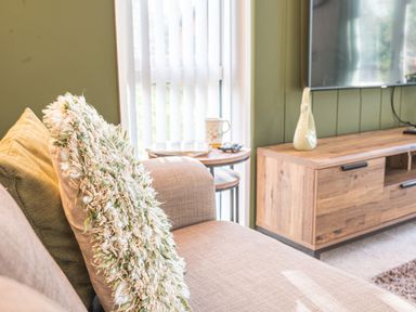 A beige sofa with textured and plain pillows next to a round side table and a wooden media unit in a living room at Fern Lodge in Allithwaite near Cartmel