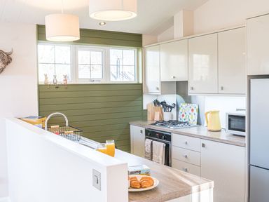 A kitchen with white cabinets a gas stove a microwave two glasses of orange juice and a plate of croissants at Fern Lodge in Allithwaite near Cartmel