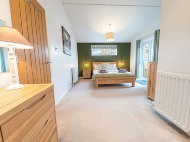 A bedroom with a wooden bed and bedside tables a wooden dresser a lamp and curtains at Fern Lodge in Allithwaite near Cartmel