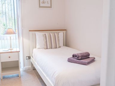 A bedroom with a single bed with white bedding and purple towels on top a nightstand with a lamp beside a window at Fern Lodge in Allithwaite near Cartmel