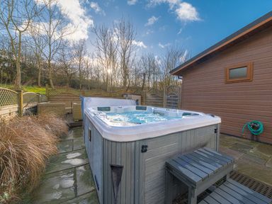 An outdoor hot tub with bubbling water on a stone patio near a wooden building and seating area at Fern Lodge in Allithwaite near Cartmel