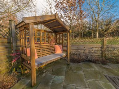 A wooden garden swing with cushions on a paved patio surrounded by wooden fencing and trees at Fern Lodge in Allithwaite near Cartmel