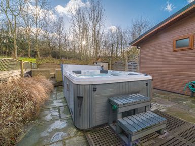 An outdoor hot tub with steps next to a wooden building and a fenced seating area with trees in the background at Fern Lodge in Allithwaite near Cartmel