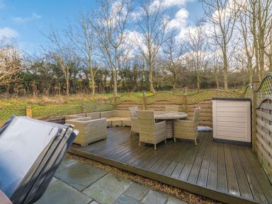 An outdoor wooden deck with rattan furniture including a round table with chairs and an L-shaped sofa at Fern Lodge in Allithwaite near Cartmel