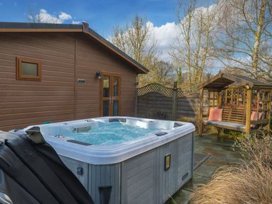 An outdoor patio with a hot tub next to a wooden cabin and a wooden covered bench with cushions at Fern Lodge in Allithwaite near Cartmel