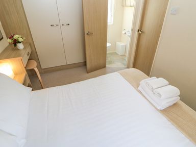A bedroom with a bed and folded towels on it a wardrobe a vanity with flowers and a partially open door leading to a bathroom at 46 Willowburn in Felton
