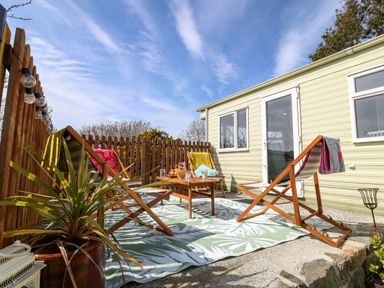An outdoor patio area with wooden deck chairs around a small wooden table on a patterned rug next to a light green mobile home at Cwrt y Ceffyl Boduan near Morfa Nefyn