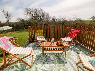 An outdoor seating area with four wooden deck chairs around a wooden table with fruit and glasses on a patterned outdoor rug