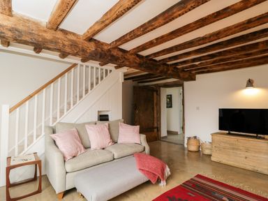 A living room with a sofa with pink cushions an ottoman with a red blanket a wooden TV unit with a television under exposed wooden ceiling beams at High Cogges Farm Holiday Cottages Witney
