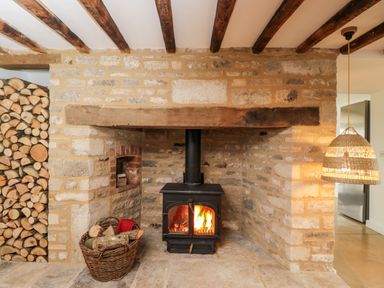A stone fireplace with a wood burning stove a basket of firewood stacked logs and a hanging woven lamp at High Cogges Farm Holiday Cottages Witney