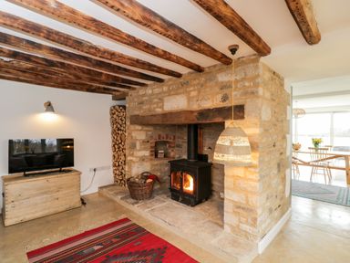 A living room with a stone fireplace and wood burner next to a TV on a wooden stand and a dining area visible through an open wall at High Cogges Farm Holiday Cottages in Witney