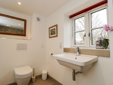 A bathroom with a toilet, wall-mounted sink, window with red blinds, mirror, and framed picture at High Cogges Farm Holiday Cottages Witney