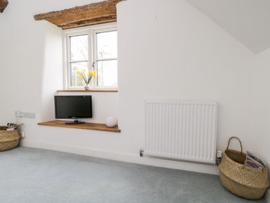 A room corner with a window, a small TV on a wooden shelf, a radiator, and woven baskets on a carpeted floor at High Cogges Farm Holiday Cottages Witney