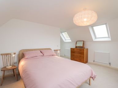 A bedroom with a double bed covered in pink bedding near two wooden chairs with lamps and a wooden dresser underneath skylight windows at High Cogges Farm Holiday Cottages Witney