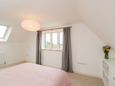 A bedroom with a bed with pink bedding grey curtains a ceiling light and flowers on a shelf at High Cogges Farm Holiday Cottages Witney
