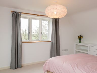 A bedroom with a window with curtains a bed with a pink duvet and a white shelf with flowers at High Cogges Farm Holiday Cottages Witney