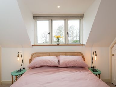 A bedroom with a bed with pink bedding two side tables with lamps and a window with flowers on the sill at High Cogges Farm Holiday Cottages Witney
