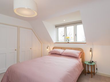 A bedroom with a bed covered in pink bedding two bedside tables with lamps and a window with flowers on the sill at High Cogges Farm Holiday Cottages in Witney