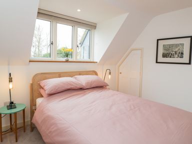 A bedroom with a double bed with pink bedding a small green side table with a lamp a window with flowers and a framed picture on the wall at High Cogges Farm Holiday Cottages in Witney