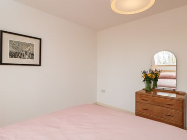 A bedroom with a pink bedspread a wooden dresser with a mirror and flowers on it and a framed picture on the wall at High Cogges Farm Holiday Cottages in Witney